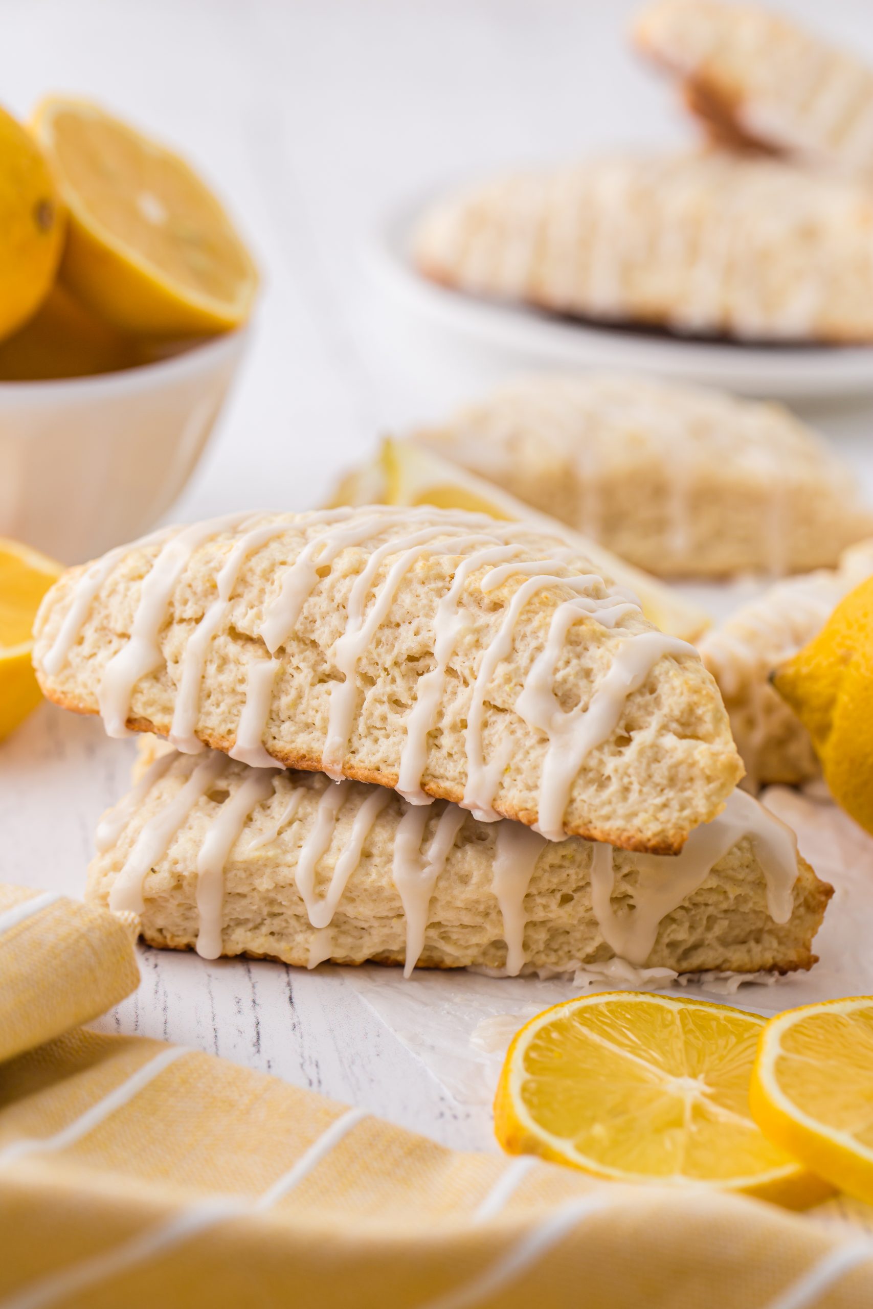 two lemon scones with glaze, stacked on each other. They're surrounded by fresh lemons. 