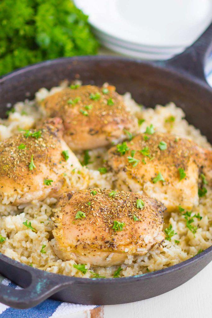 Seasoned baked chicken thighs rest atop garlic parmesan rice in a black cast iron skillet, creating an irresistible one pan chicken meal garnished with fresh parsley. Plates and green herbs are blurred in the background.