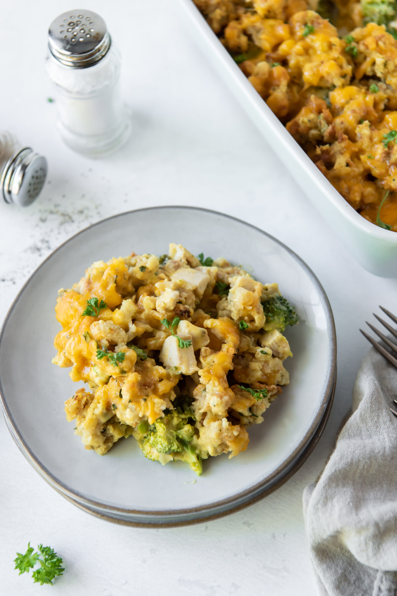 An overhead view of a portion of chicken broccoli stuffing casserole on a white plate. A casserole dish rests next to it. 