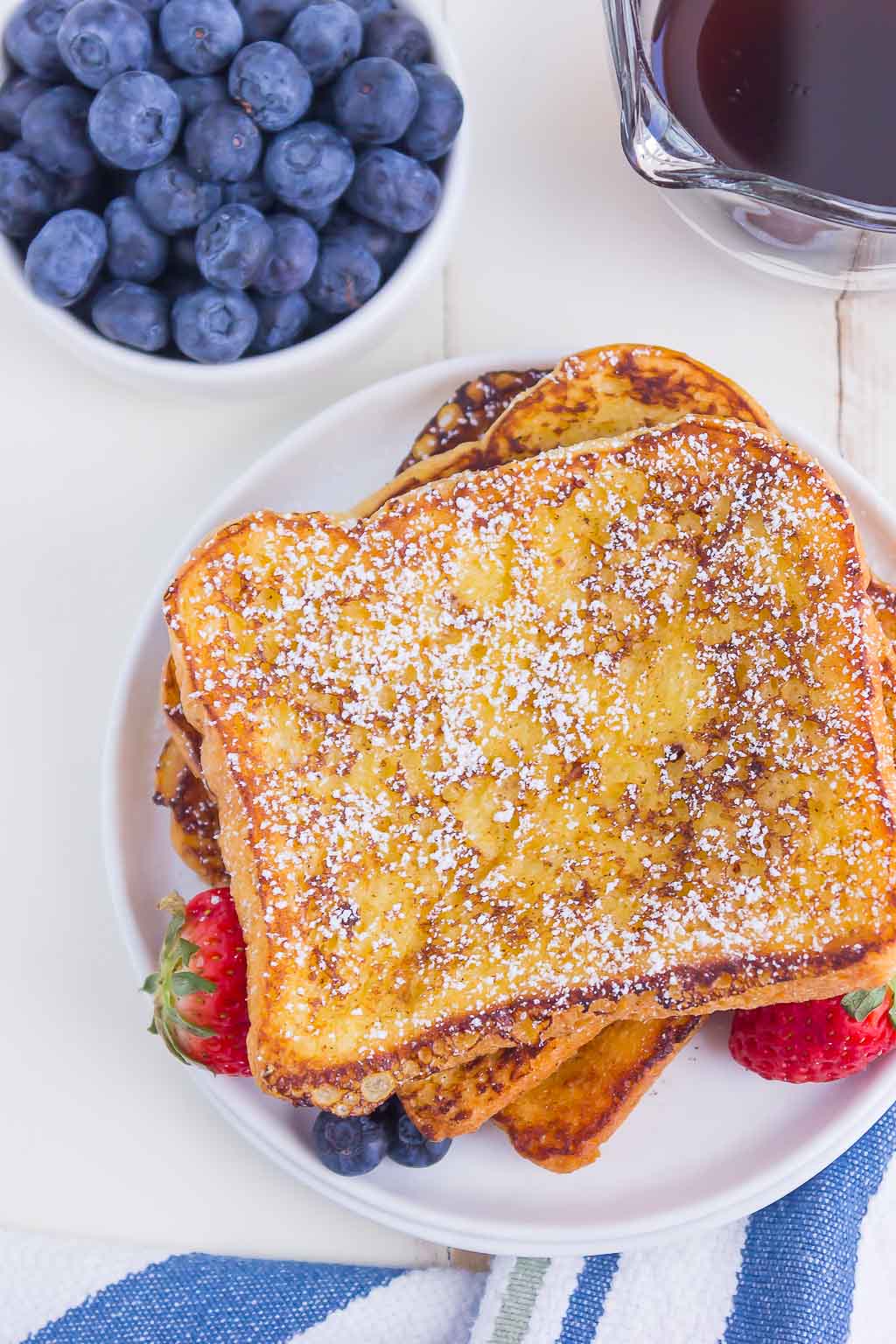 An overhead view of a stack of brioche bread french toast. A small dish of blueberries and a bowl of syrup rest nearby. 