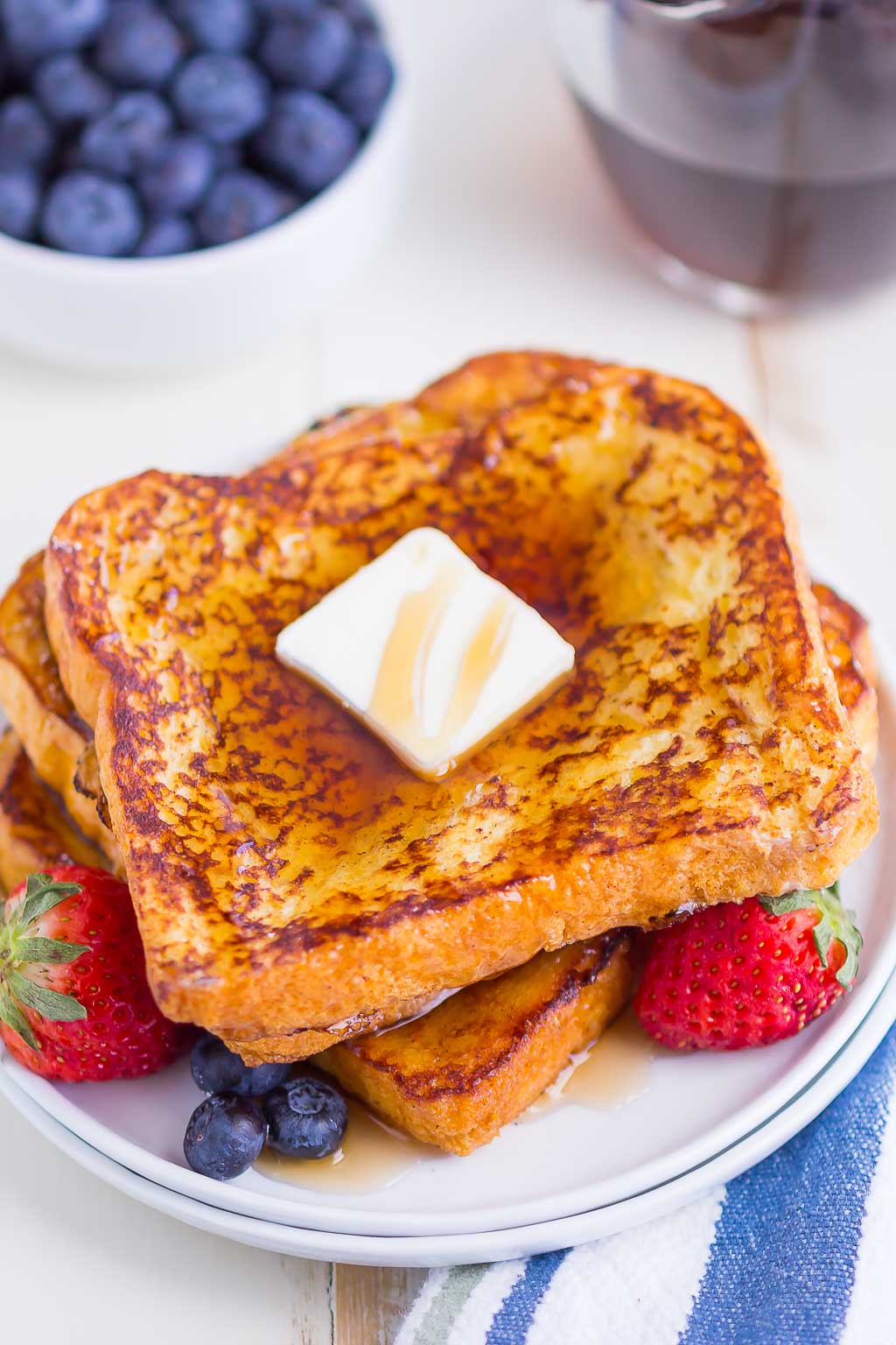 Three slices of brioche french toast stacked on a small white plate. The stack is topped with butter and maple syrup, and there are fresh berries on the plate. 