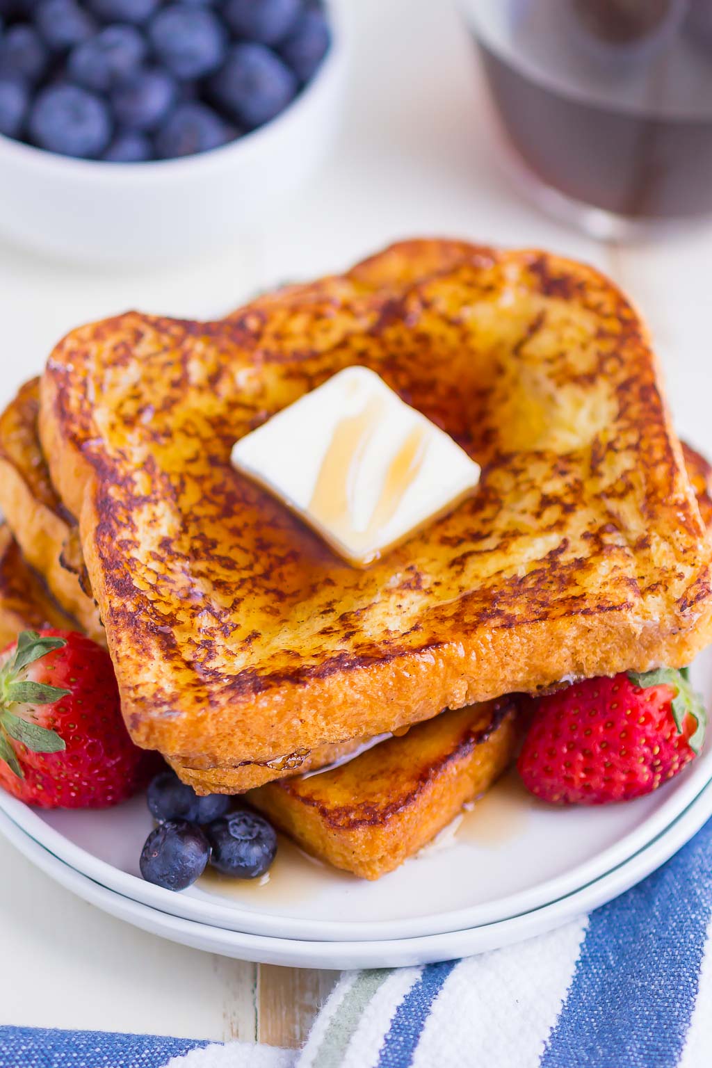 Three slices of brioche bread french toast stacked on a small white plate. The stack is topped with butter and maple syrup, and there are fresh berries on the plate. 