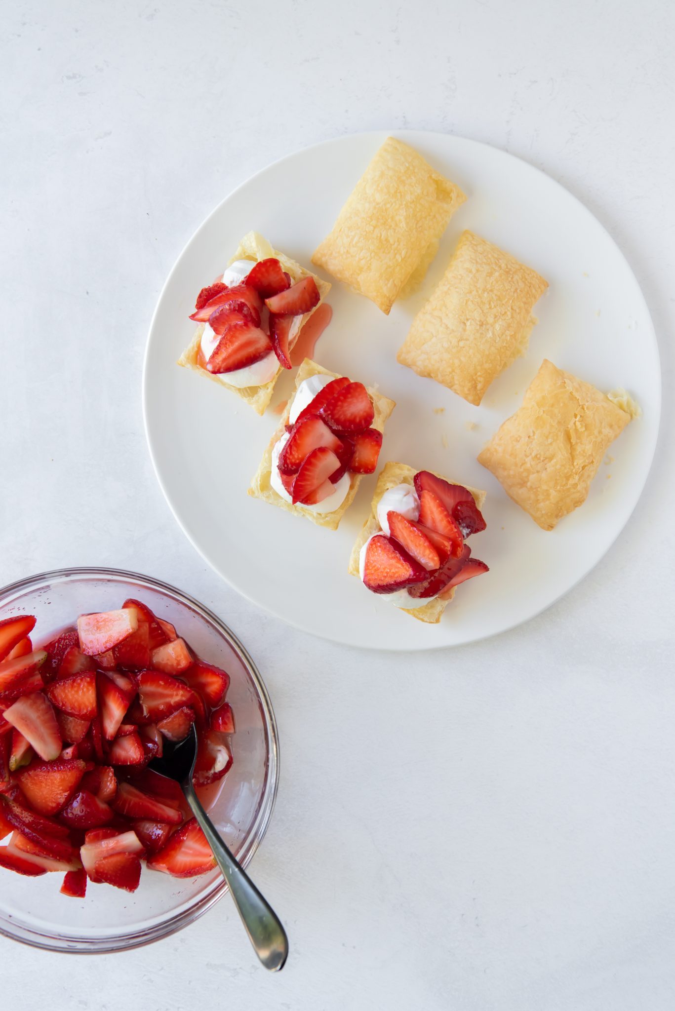 A white plate with squares of easy strawberry shortcake puffs, some topped with whipped cream and sliced strawberries, sits next to a glass bowl filled with sliced strawberries and a spoon.
