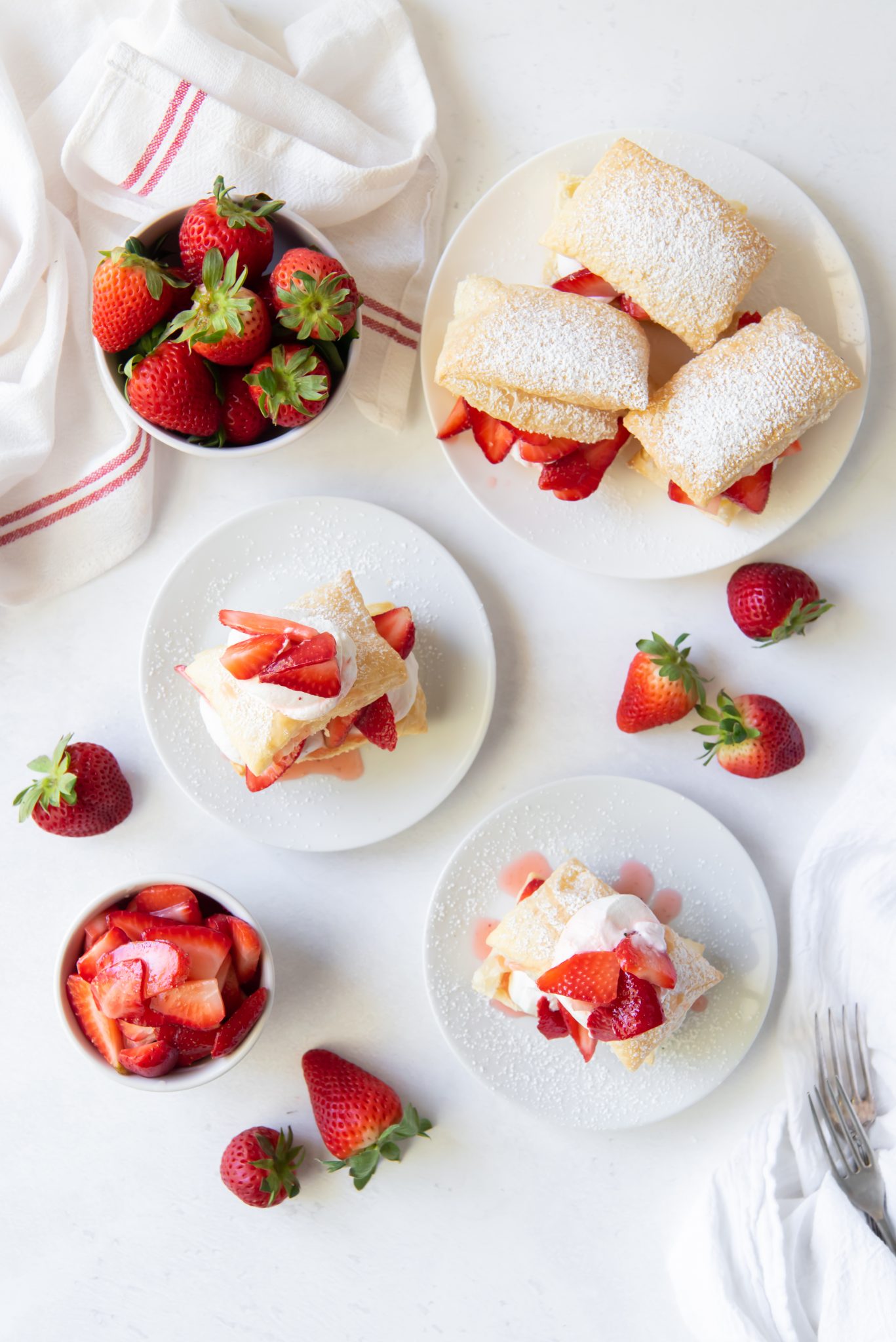 Plates of easy strawberry shortcake puffs with whipped cream and fresh strawberries sit on a white table, surrounded by whole strawberries, a bowl of sliced strawberries, a striped cloth, and silver forks.