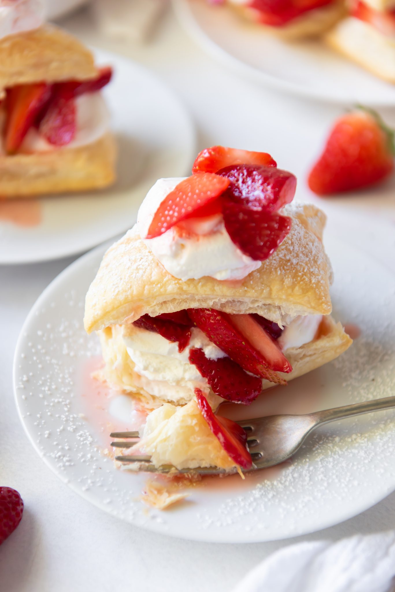 A close-up of easy strawberry shortcake puffs featuring flaky pastry layers, whipped cream, and fresh strawberry slices on a white plate. A fork rests nearby, and powdered sugar is sprinkled on top.