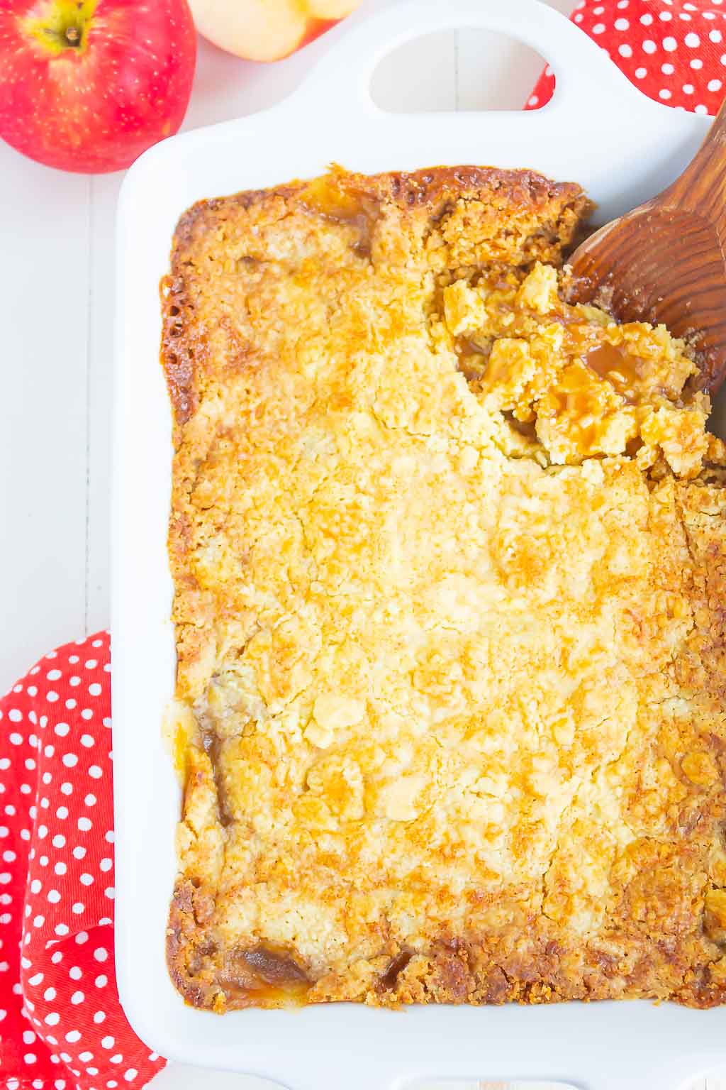 An overhead view of an apple dump cake in a white baking dish. A wooden spoon is scooping out some of the cake. 