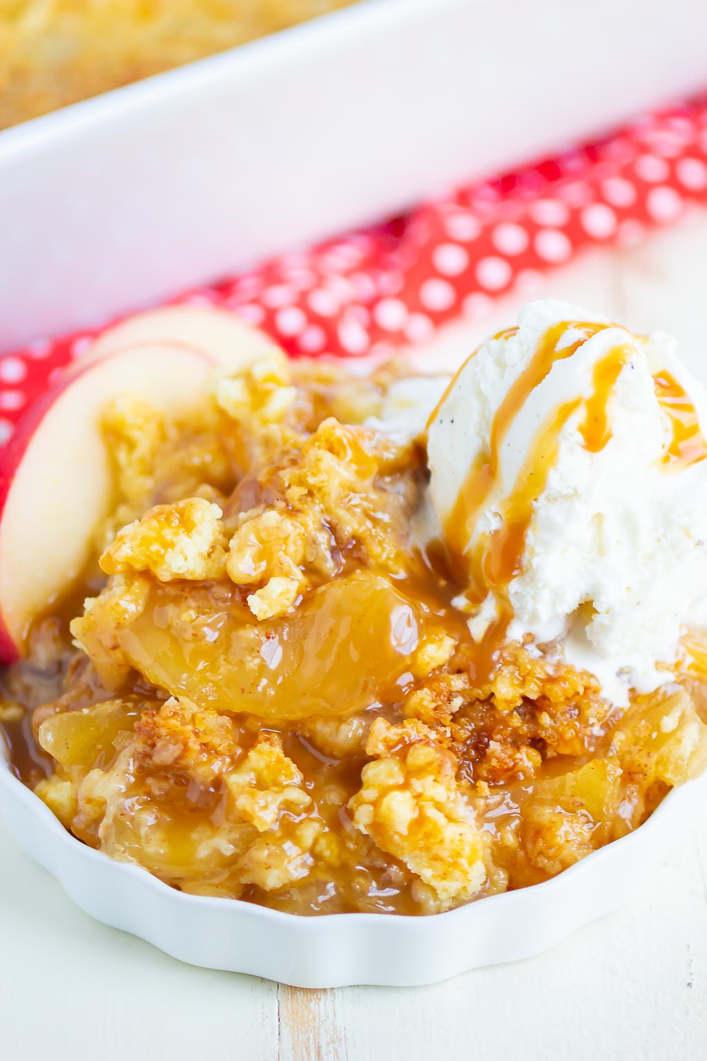 A portion of caramel apple dump cake in a white ramekin. Two apple slices and a scoop of ice cream garnish the dish. 