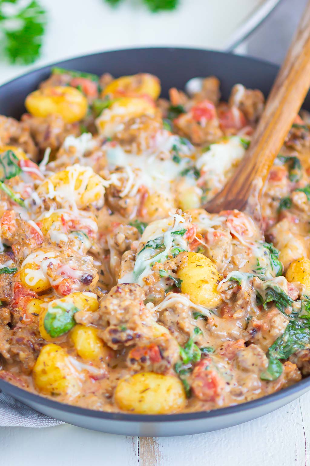 A close up view of a skillet of gnocchi with sausage and spinach, with a wooden serving spoon. 