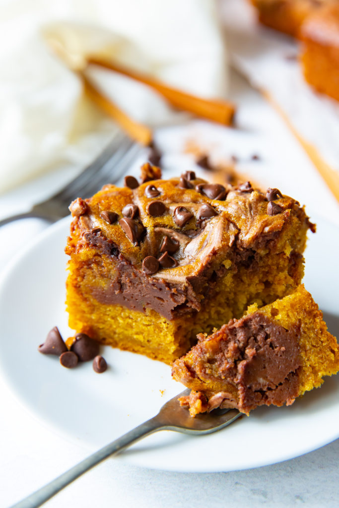 A slice of pumpkin chocolate bar with chocolate swirls sits on a white plate, alongside pumpkin cream cheese bars. A fork holds a bite-sized piece in the foreground, with chocolate chips sprinkled on top and cinnamon sticks in the background.