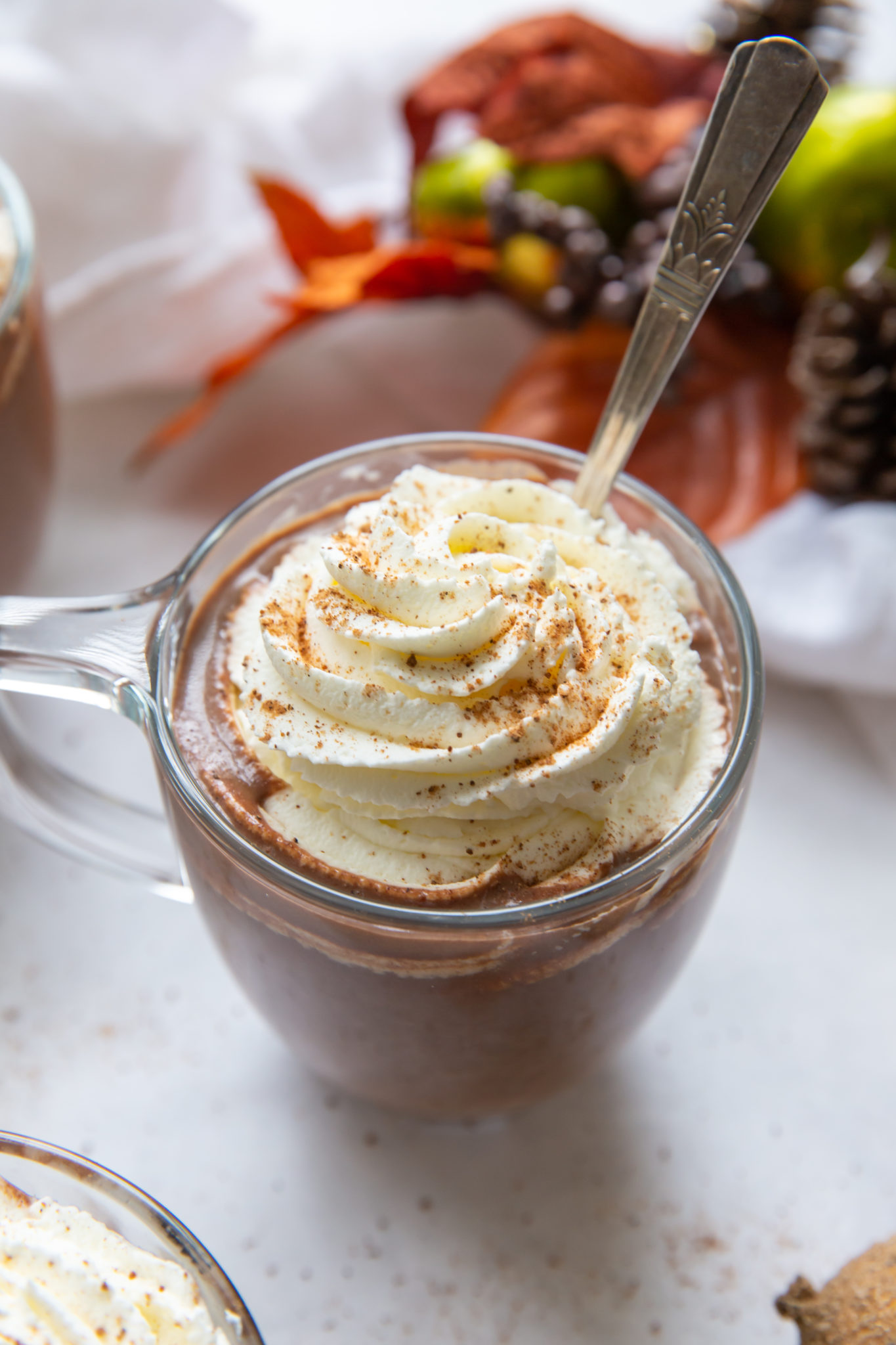 A glass mug of pumpkin spice hot chocolate garnished with whipped cream. A spoon is sticking out of the mug.