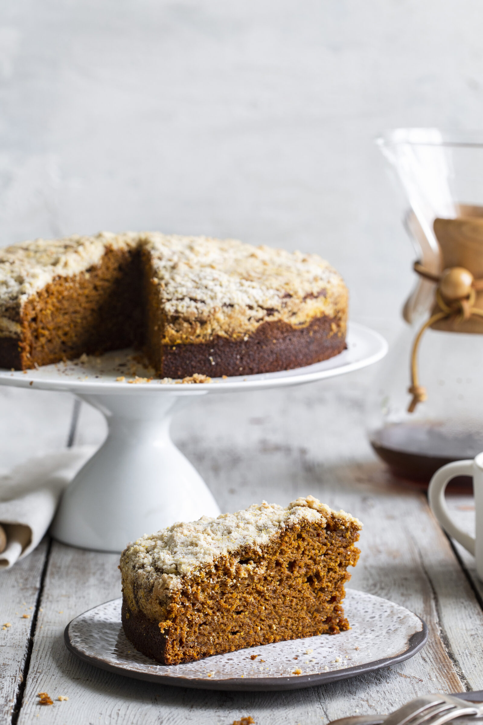 A pumpkin spice coffee cake on a white cake stand, with a slice on a dessert plate in the foreground. 