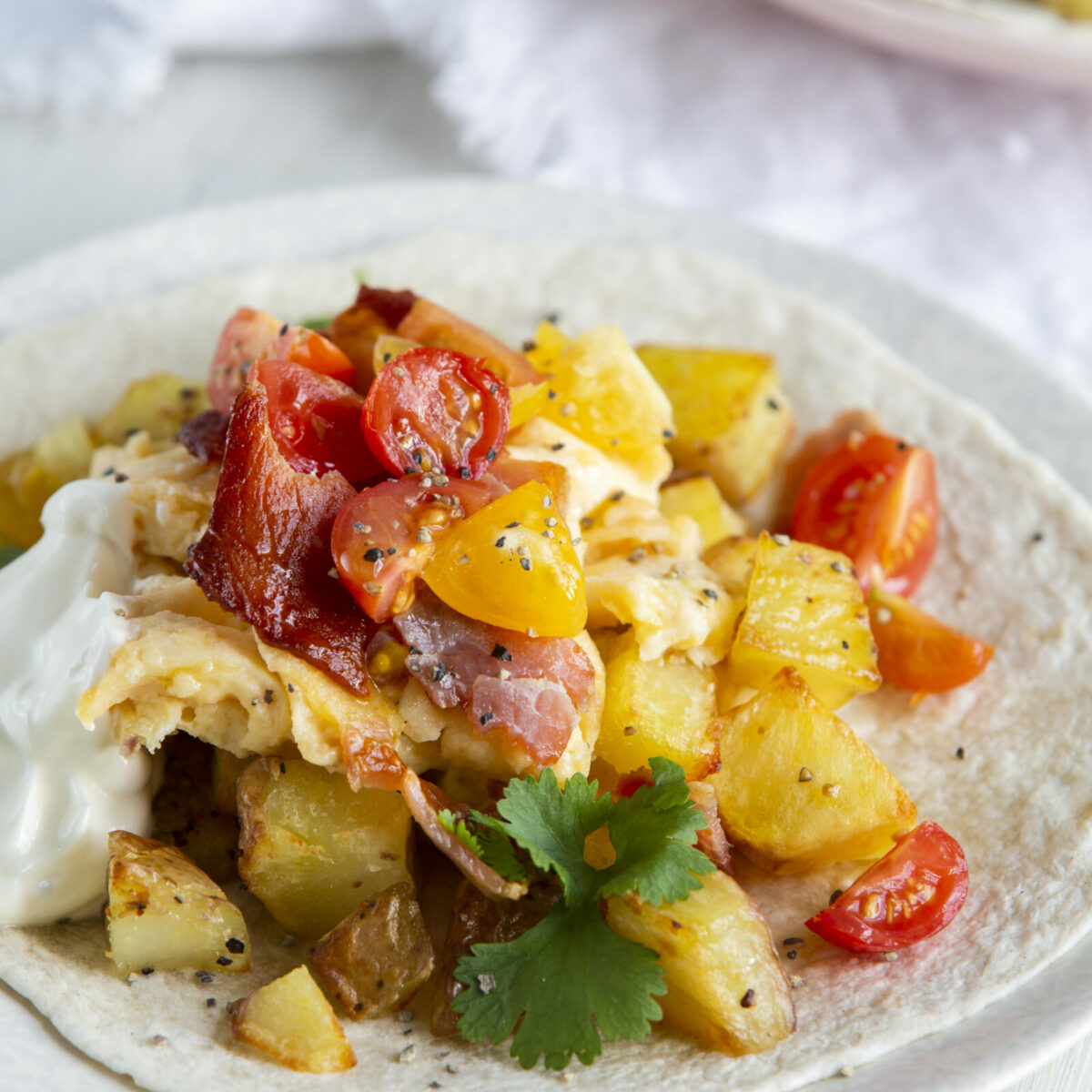 Enjoy easy breakfast tacos with a soft tortilla topped with scrambled eggs, roasted potatoes, cherry tomatoes, bacon, a dollop of sour cream, and a sprig of cilantro, served on a white plate.