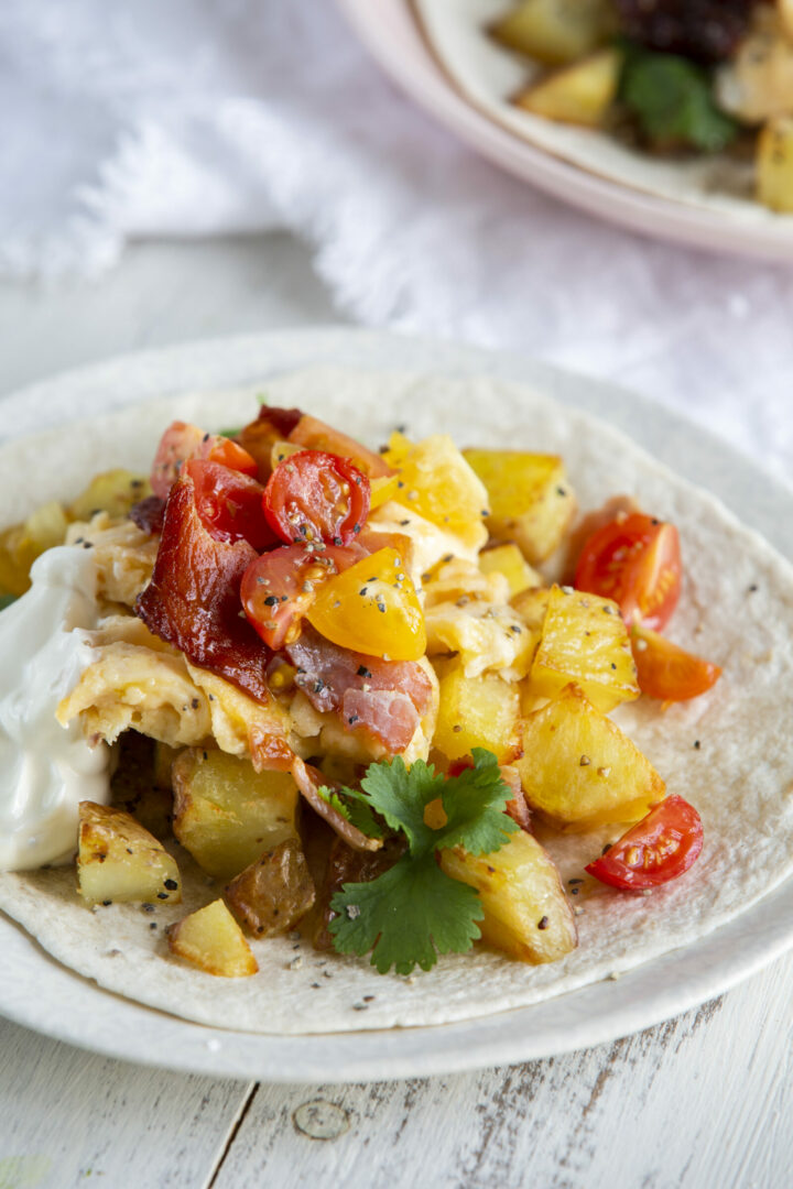 Enjoy easy breakfast tacos with a soft tortilla topped with scrambled eggs, roasted potatoes, cherry tomatoes, bacon, a dollop of sour cream, and a sprig of cilantro, served on a white plate.