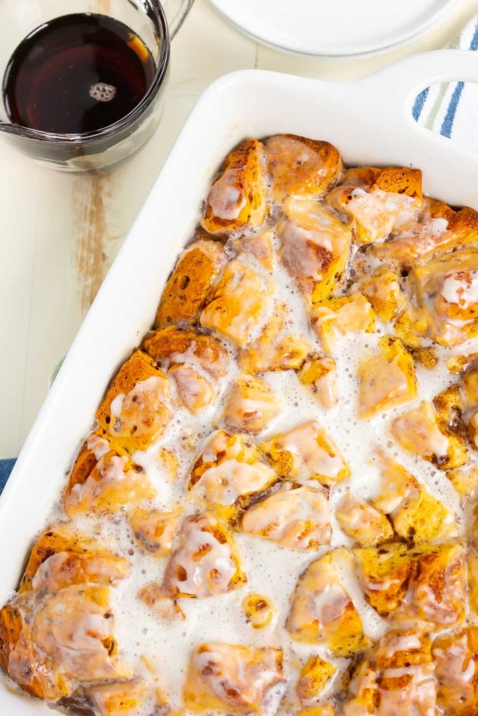 A white baking dish filled with cinnamon roll French toast casserole sits beside a glass mug of coffee and stacked white plates on a light-colored table.