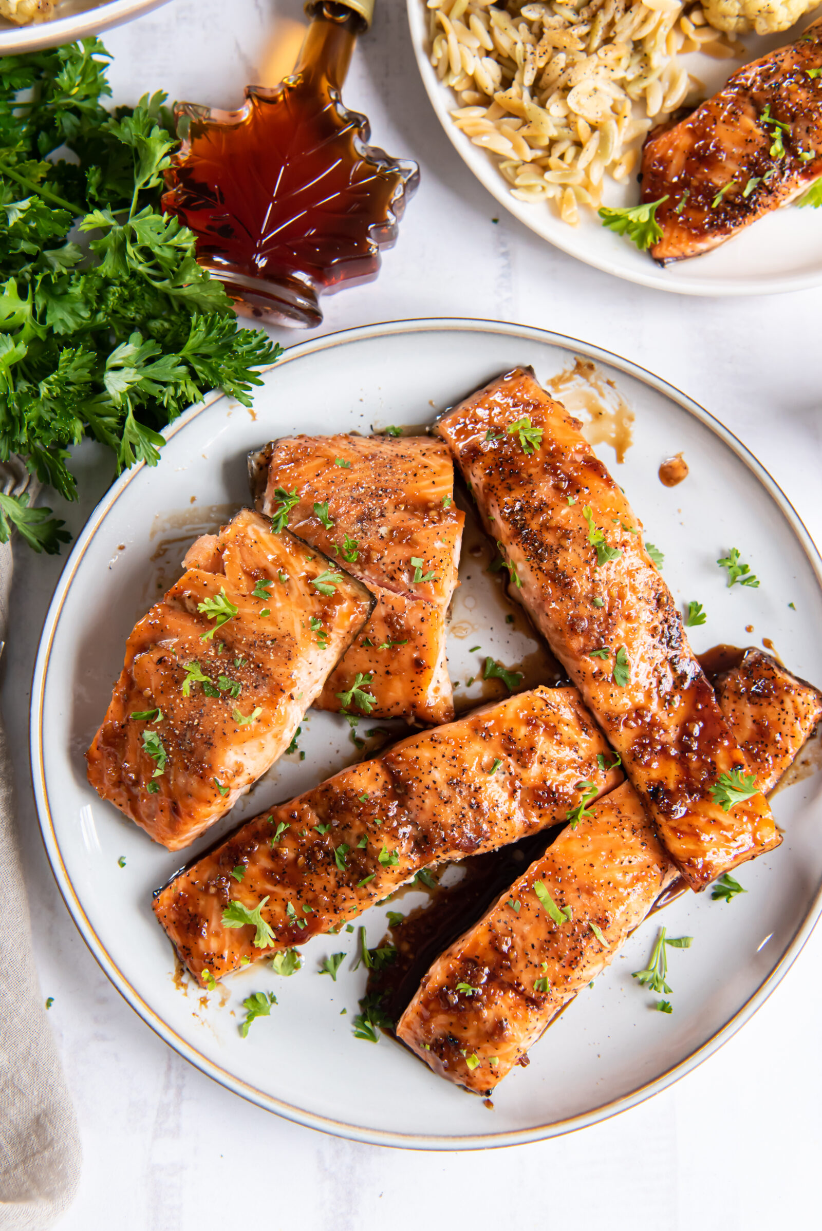 Overhead view of five filets of maple syrup soy sauce salmon on a white plate. A bottle of maple syrup and a bunch of parsley rest on the side. 