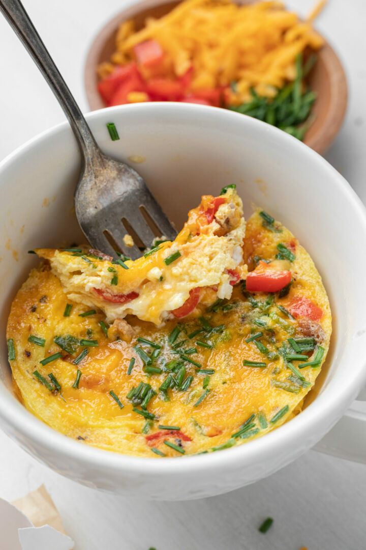 A fork lifts a bite of a microwave mug omelet with chopped chives, tomatoes, and melted cheese in a white mug. In the background, a small bowl holds extra shredded cheese, chives, and tomato pieces.