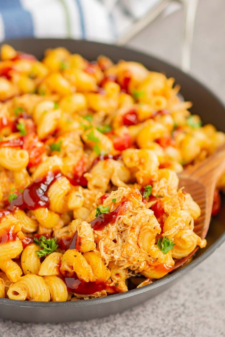 A close-up of a skillet filled with cheesy macaroni pasta, shredded chicken, barbecue sauce, and fresh parsley—a delicious one pot BBQ chicken pasta recipe. A wooden spoon is scooping up a serving.