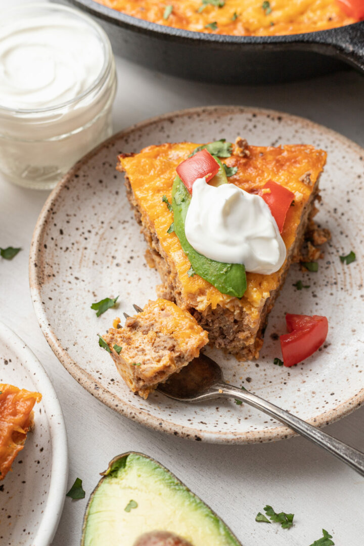 A slice of taco egg bake casserole topped with sliced avocado, diced tomato, and a dollop of sour cream sits on a speckled plate, with a fork taking a bite. A jar of sour cream and half an avocado are nearby.
