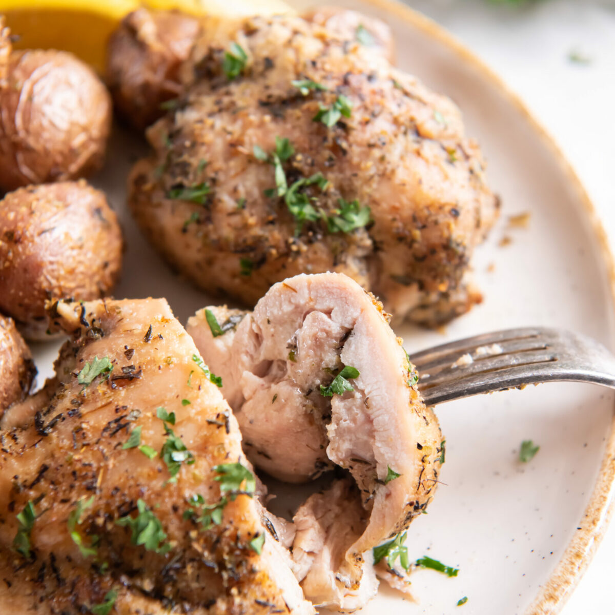 A close-up of air fryer chicken thighs and potatoes, seasoned and baked to perfection, garnished with chopped herbs on a plate with lemon wedges, as a fork cuts into the juicy chicken.