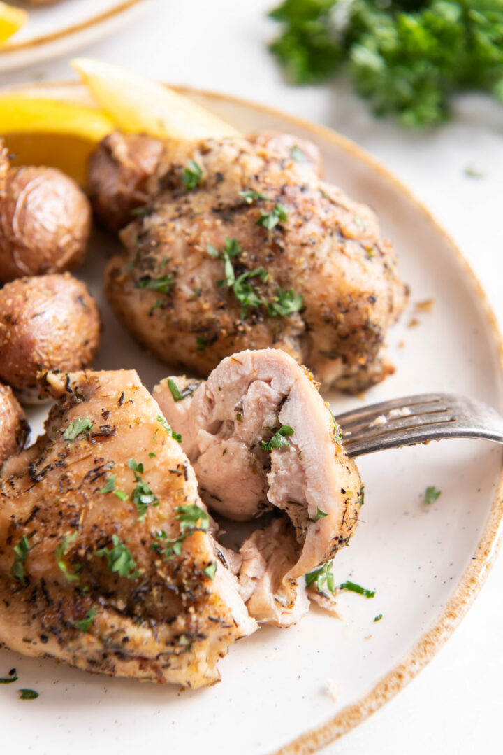 A close-up of air fryer chicken thighs and potatoes, seasoned and baked to perfection, garnished with chopped herbs on a plate with lemon wedges, as a fork cuts into the juicy chicken.