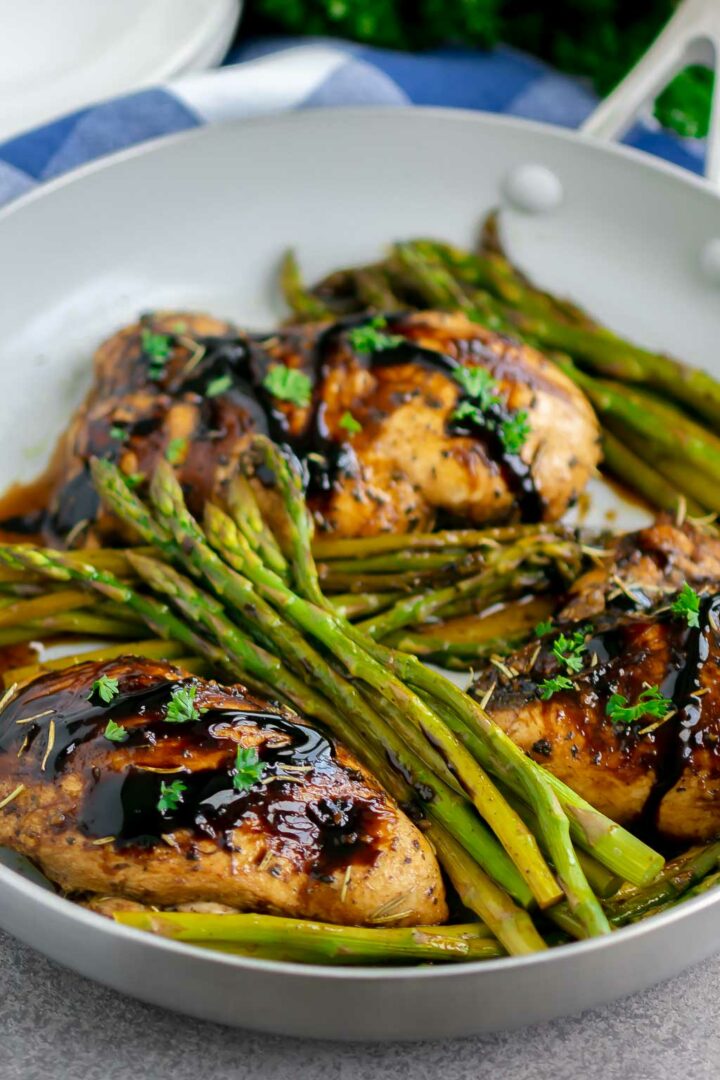 Three cooked chicken breasts with a dark balsamic glaze and fresh herbs, arranged in a skillet with roasted asparagus spears. This one pan balsamic chicken and asparagus dish is served alongside plates and a cloth napkin in the background.