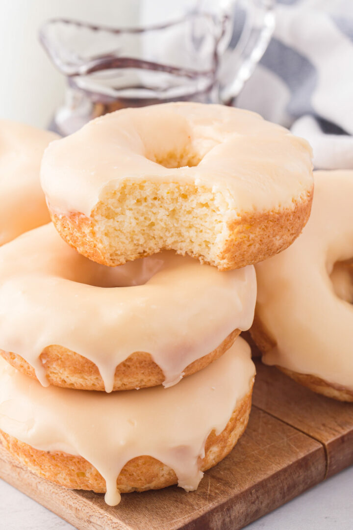 A stack of three glazed maple donuts sits on a wooden board, topped with a bitten donut. In the background, a glass container of chocolate sauce is softly blurred.