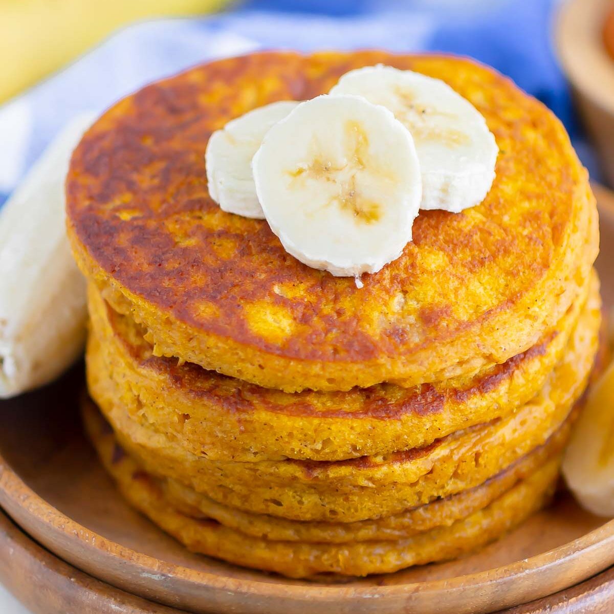 a stack of pumpkin banana pancakes, drizzled with maple syrup and topped with banana slices, on a wooden plate