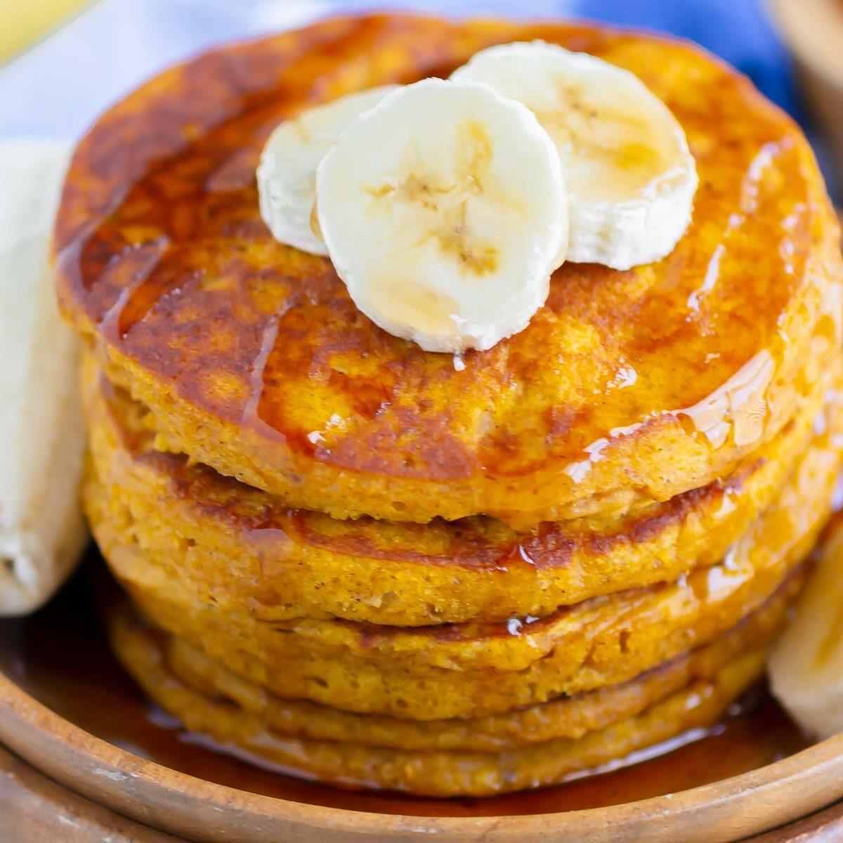 a stack of pumpkin banana pancakes, drizzled with maple syrup and topped with banana slices, on a wooden plate