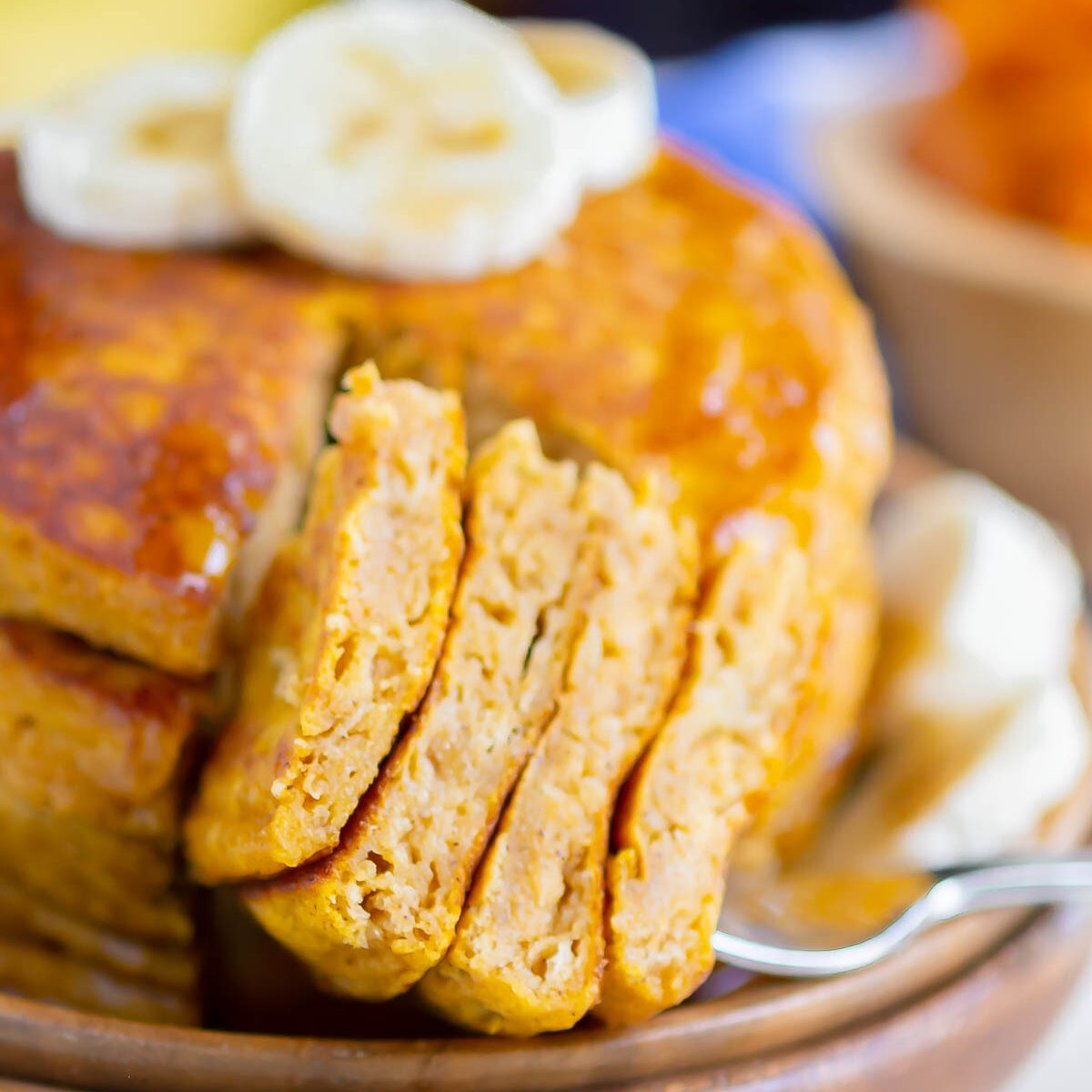 a stack of forkful pumpkin banana pancakes bites, drizzled with maple syrup and topped with banana slices, on a wooden plate