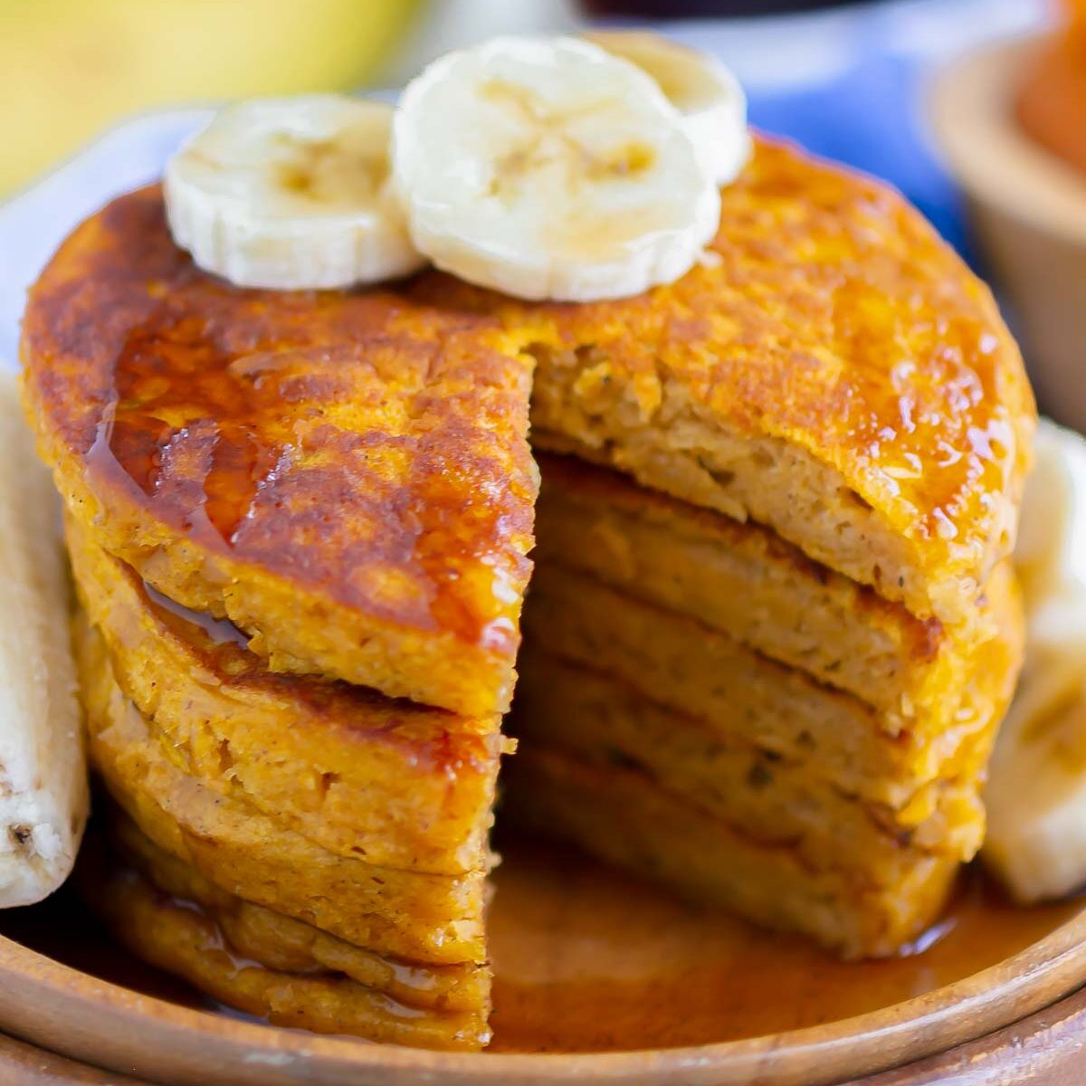 a stack of slice cut out pumpkin banana pancakes, drizzled with maple syrup and topped with banana slices, on a wooden plate