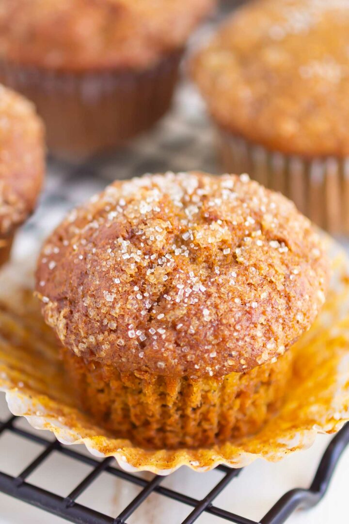 A close-up of a pumpkin banana muffin topped with coarse sugar sits on an open paper liner on a cooling rack, with more muffins blurred in the background.