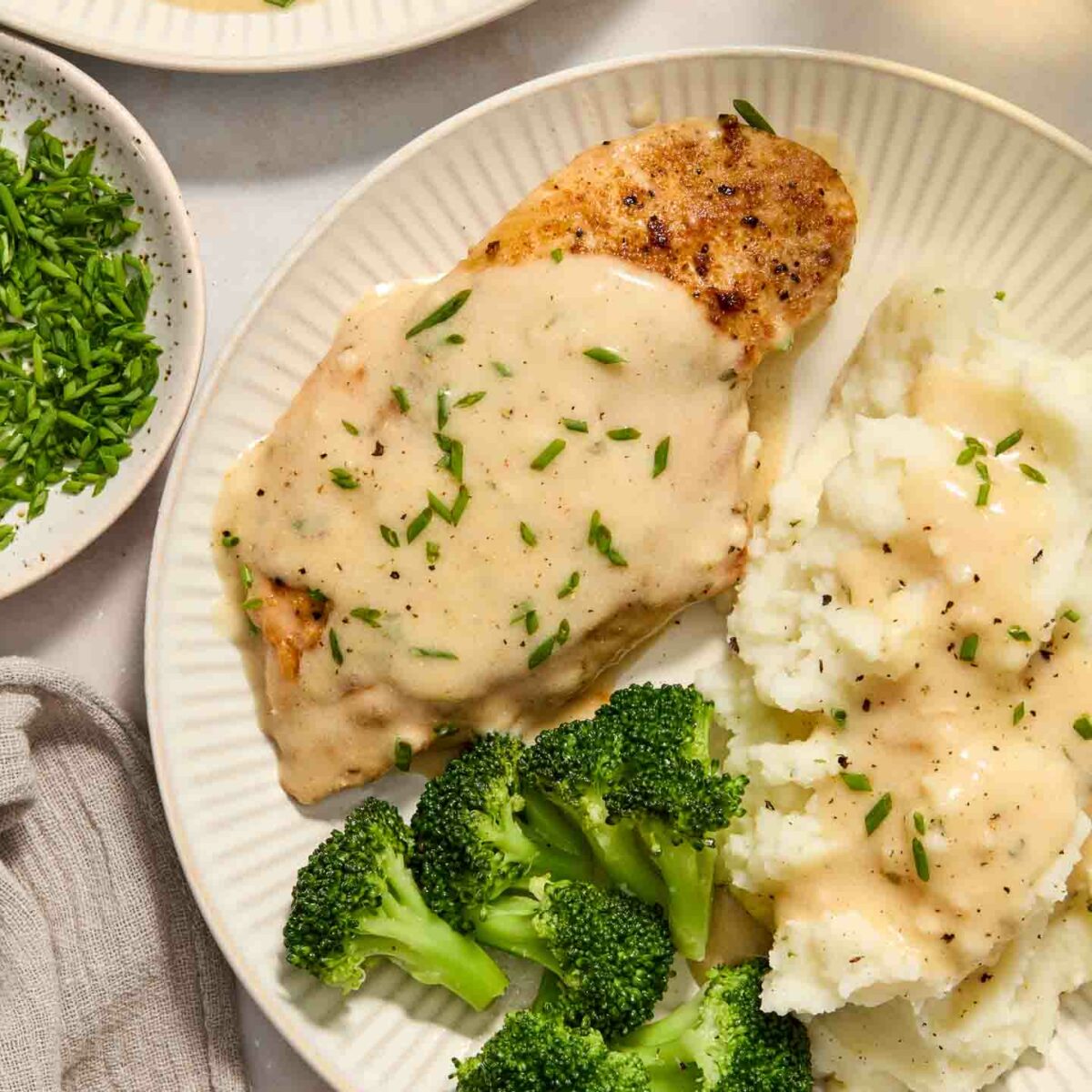 A plate of creamy ranch chicken breast topped with chopped chives, mashed potatoes with gravy, and steamed broccoli on a white plate. A bowl of chopped herbs and a glass of drink are nearby.