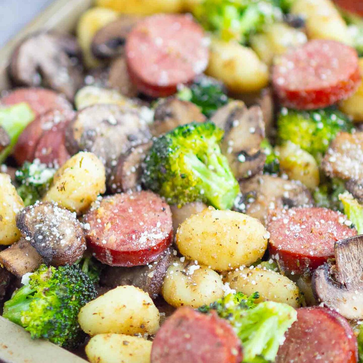 A sheet pan sausage gnocchi dinner featuring roasted gnocchi, sliced sausage, broccoli florets, and mushrooms, all sprinkled with grated cheese and black pepper.