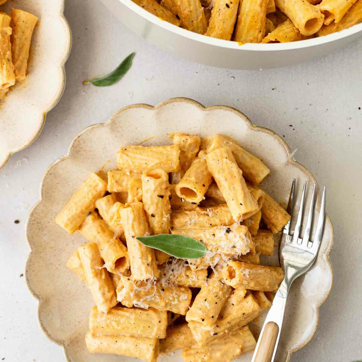 pumpkin alfredo pasta on white plate with a silver fork next to another plate of pasta and rest of pasta in white skillet