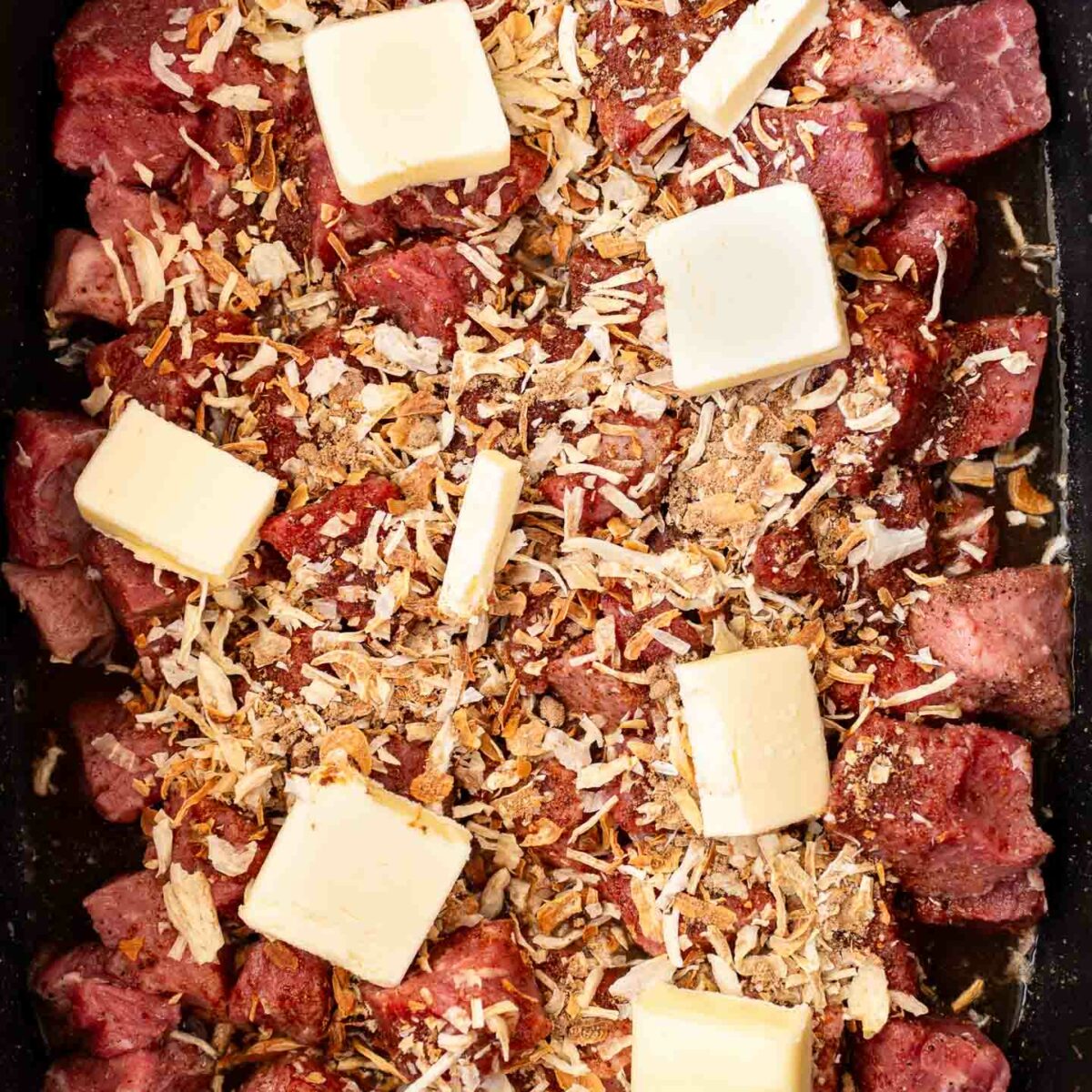 raw steak bites in the slow cooker with seasonings and sliced butter on top