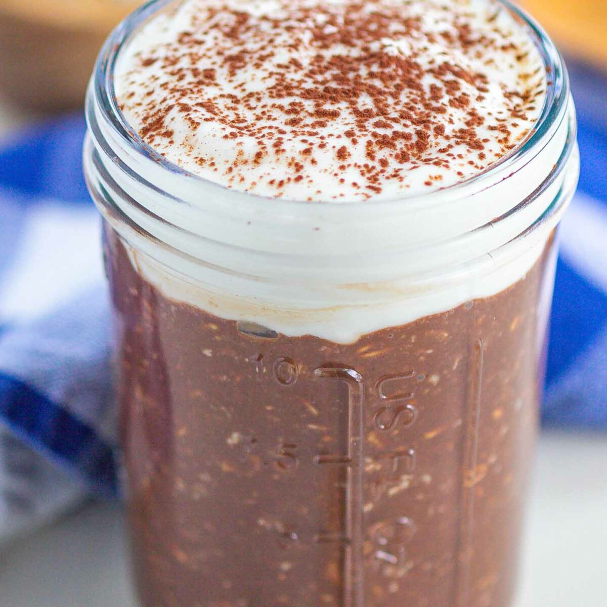 A mason jar filled with tiramisu overnight oats, topped with a layer of mascarpone yogurt and a dusting of cocoa powder, sits on a table with a blue and white cloth in the background.