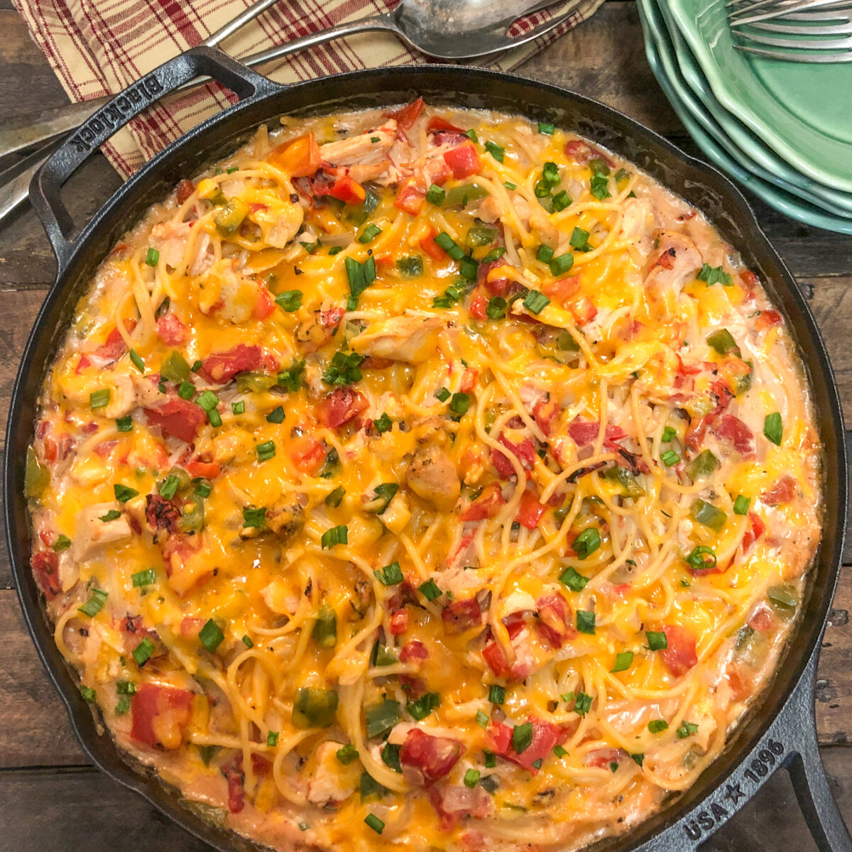 A skillet filled with baked cheesy spaghetti, featuring pasta, diced tomatoes, green onions, and melted cheese. A folded checkered napkin with utensils is beside the skillet on a rustic wooden table.