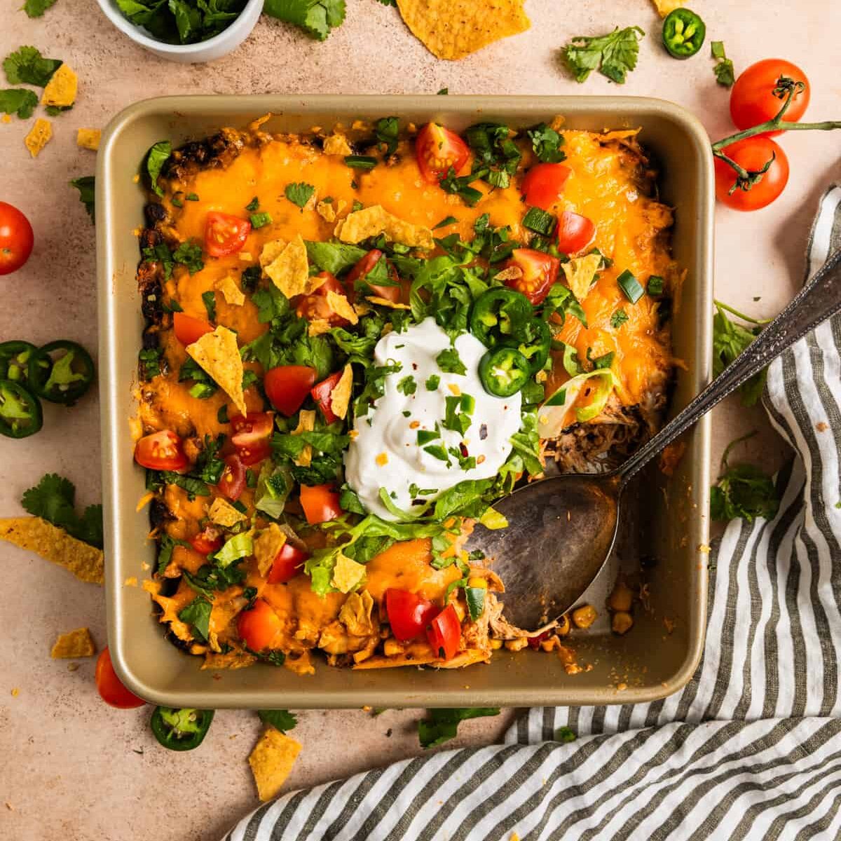 A square pan filled with a taco casserole topped with melted cheese, chopped tomatoes, jalapeños, cilantro, and a dollop of sour cream. A spoon rests inside the pan. Tortilla chips and cherry tomatoes are scattered around.