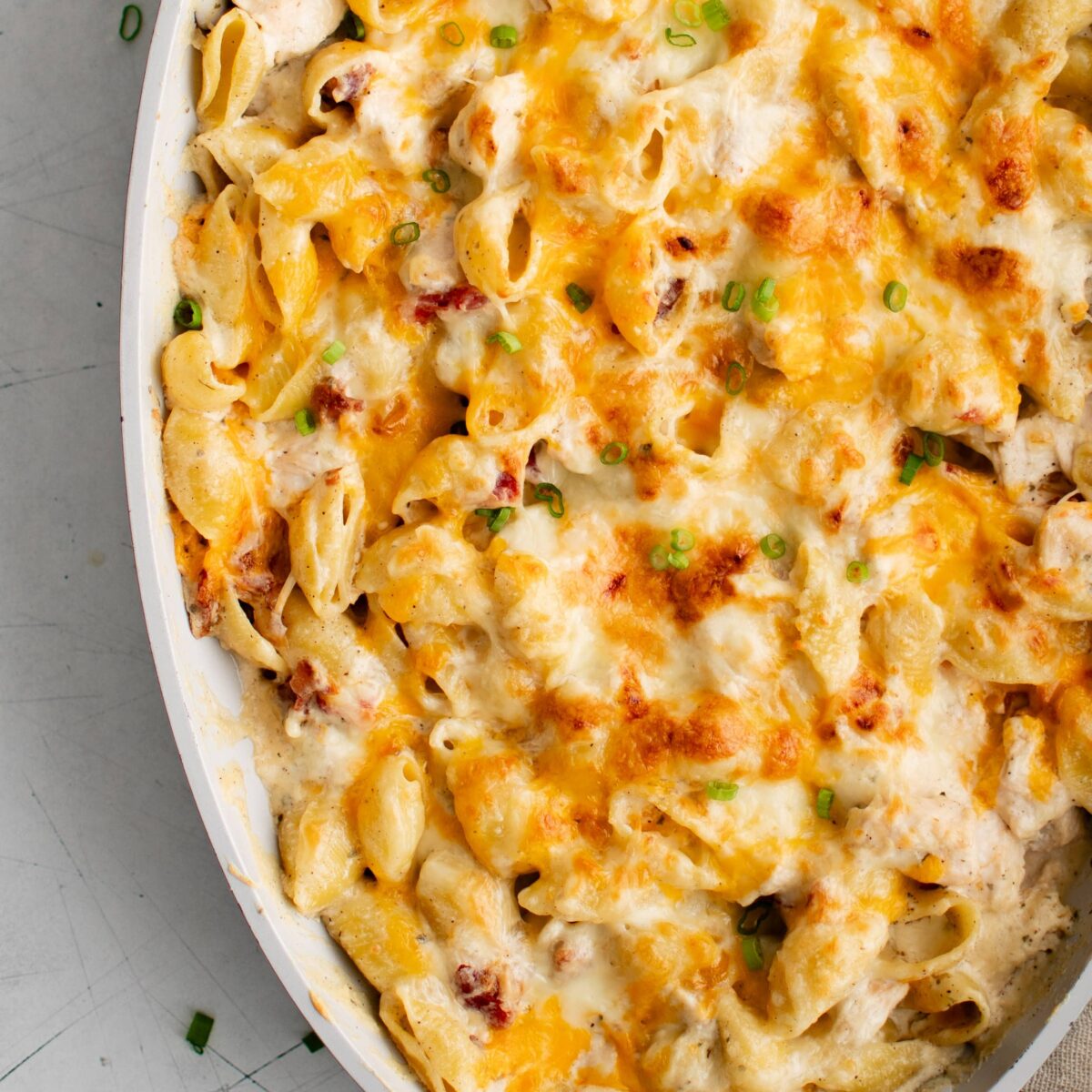 A baked pasta dish with melted cheese and browned top filling an oval white casserole dish. Small pieces of green garnish are sprinkled over the cheese. A striped cloth is partially visible underneath the dish.