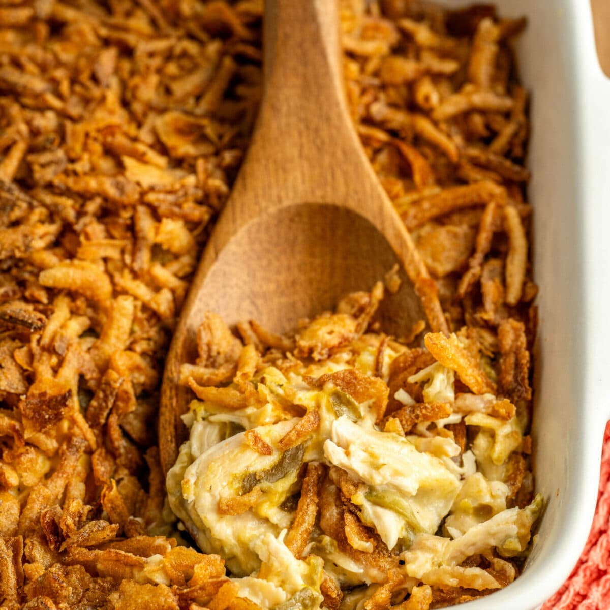A white casserole dish filled with crispy baked topping and a creamy mixture beneath. A wooden spoon rests in the dish, scooping up some of the creamy filling. The dish is placed on a red textured cloth.