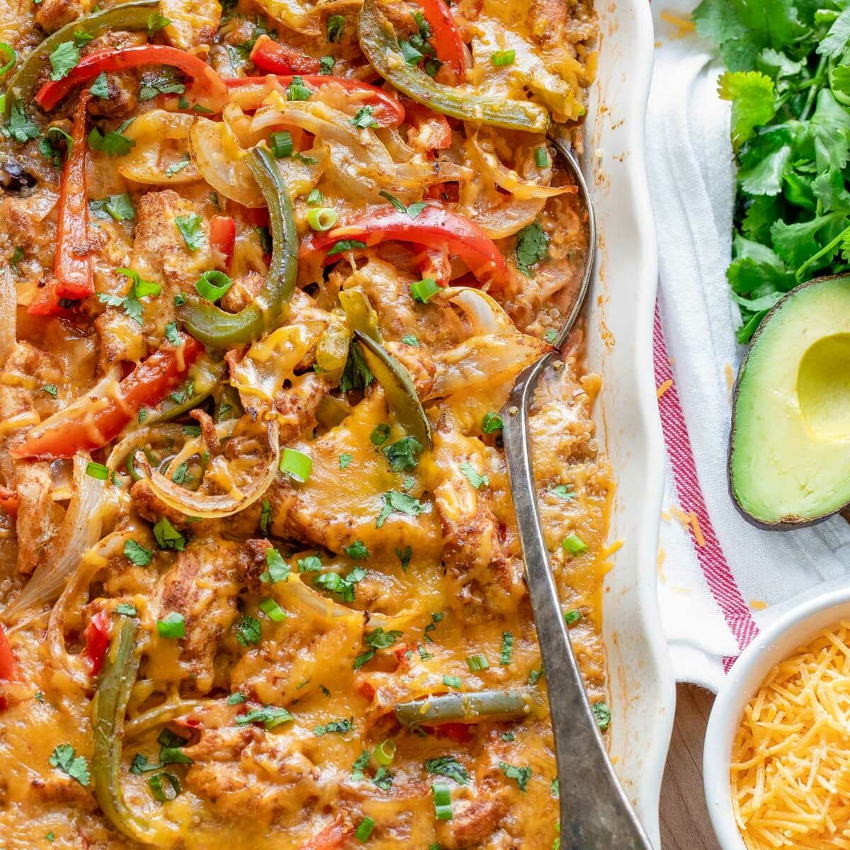A casserole dish filled with cheesy chicken and vegetable bake, including red and green bell peppers and onions. A spoon is in the dish. Next to it are chopped green onions, cilantro, sliced avocado, and shredded cheese in bowls.