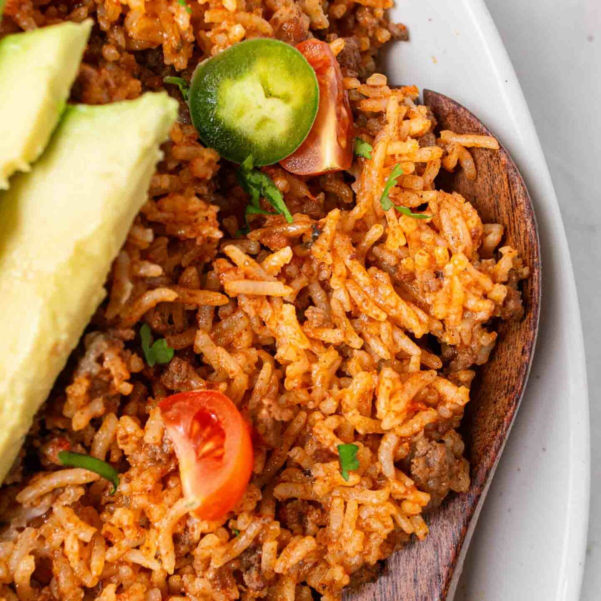 taco rice shown in a white dish with a wooden spoon and topped with sliced avocado and jalapeños