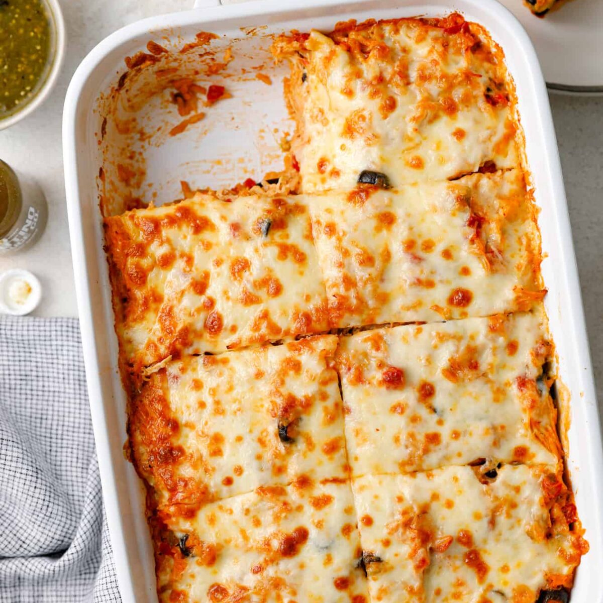 A white rectangular baking dish filled with cheesy enchiladas, topped with melted cheese and sliced into squares. A portion is missing from one corner. Next to the dish are lime wedges, a fork, and a small bowl of green sauce.