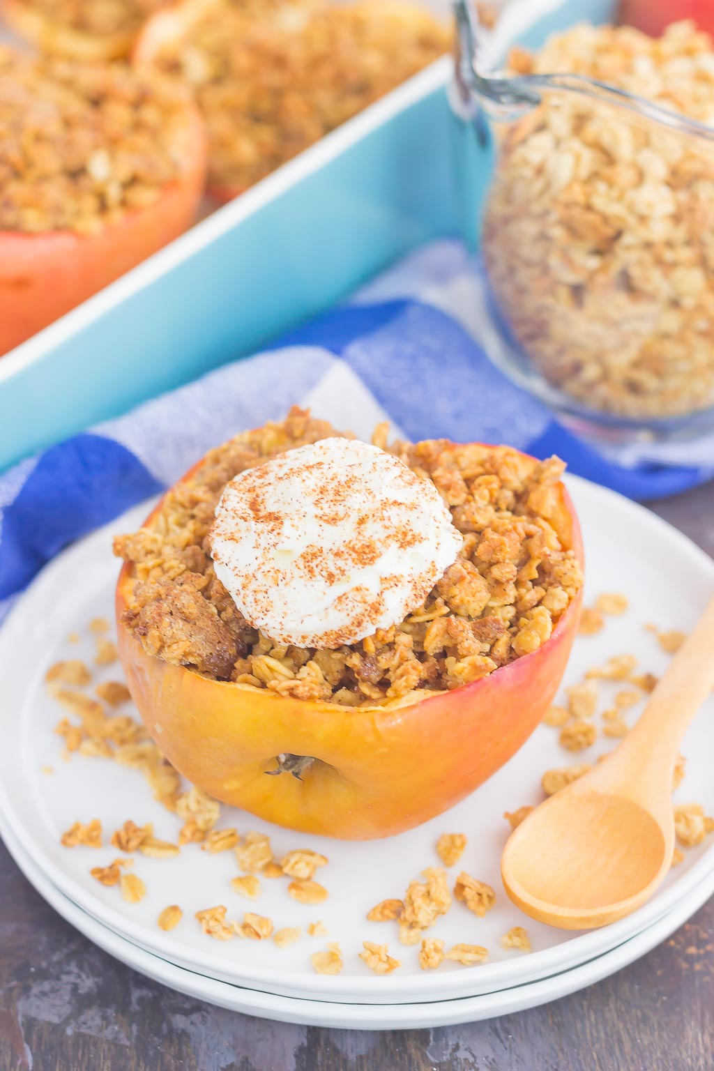 apple with granola on a white plate with a dollop of whipped cream and a wooden spoon