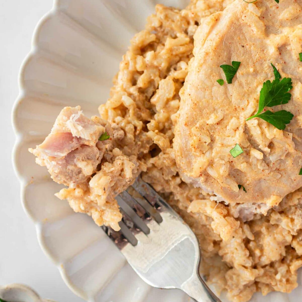 A creamy pork chop rests on a bed of rice on a scalloped white plate. Garnished with fresh parsley, a fork holds a bite-sized piece over the rice. A small dish of parsley is partially visible in the corner.
