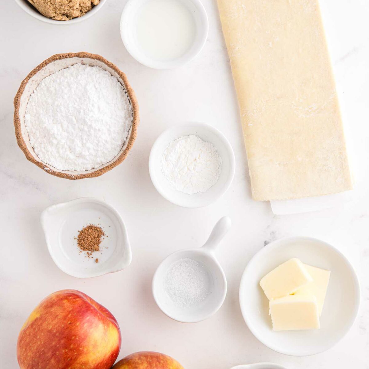 Various baking ingredients are arranged on a white surface: brown sugar, powdered sugar, dough, milk, powdered sugar, spices, butter, two apples, and small bowls with spices.