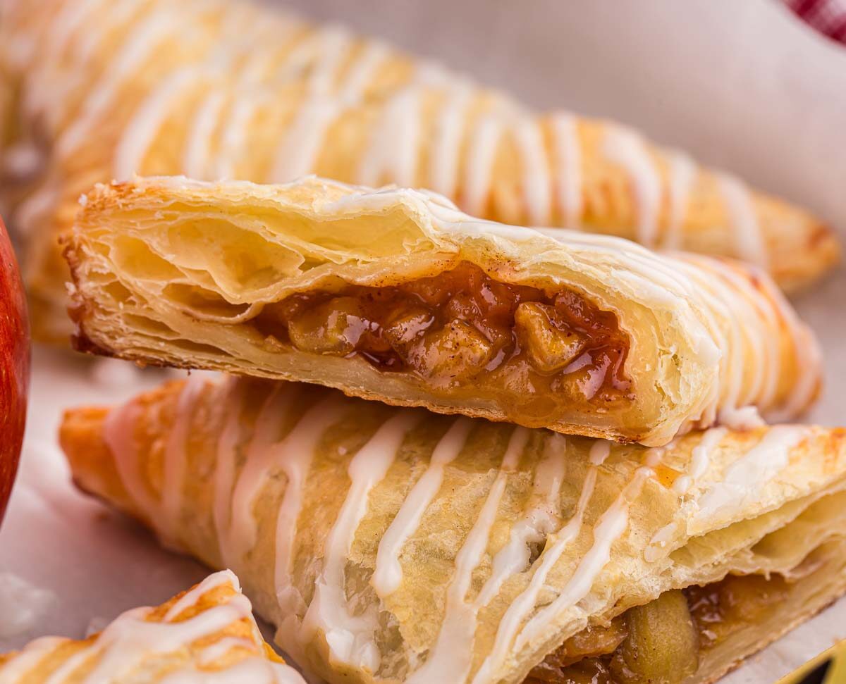 Close-up of a pastry filled with apple chunks and cinnamon, drizzled with white icing. The pastry is flaky and golden brown. A whole red apple is partially visible on the left, hinting at the pastrys filling.