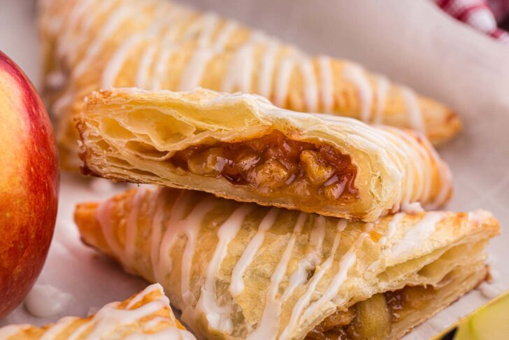 Close-up of a pastry filled with apple chunks and cinnamon, drizzled with white icing. The pastry is flaky and golden brown. A whole red apple is partially visible on the left, hinting at the pastrys filling.
