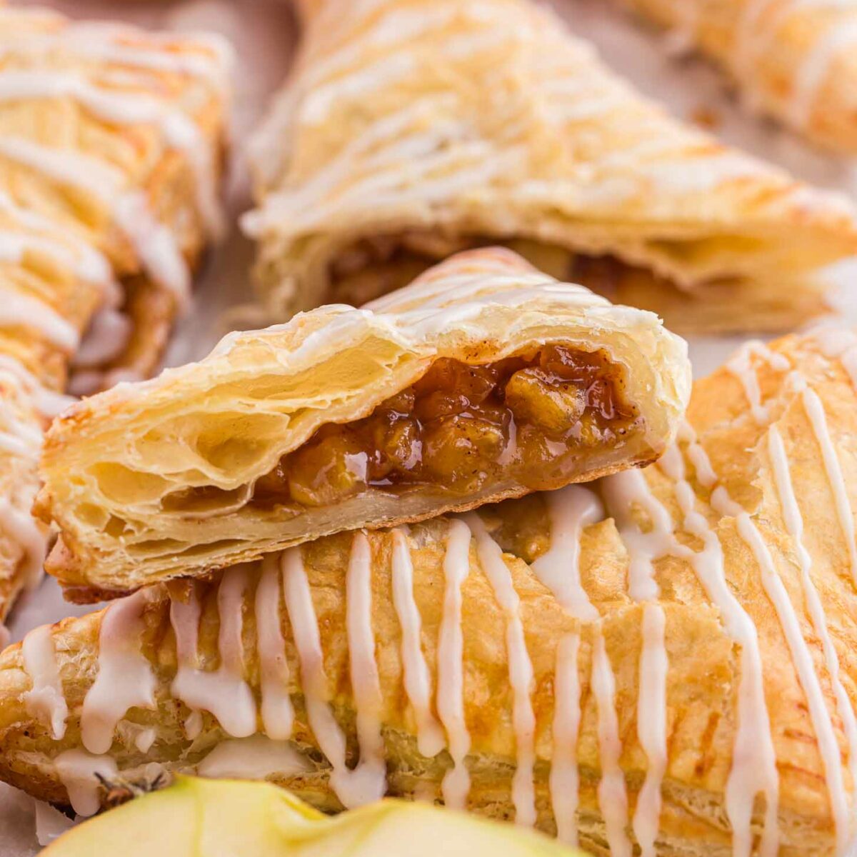 Flaky apple turnovers are drizzled with white icing. One turnover is cut open to reveal a spiced apple filling. A fresh apple is partially visible in the background.