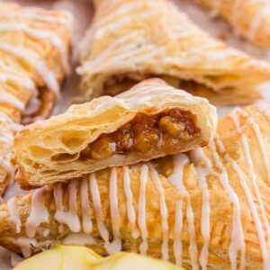 Flaky apple turnovers are drizzled with white icing. One turnover is cut open to reveal a spiced apple filling. A fresh apple is partially visible in the background.